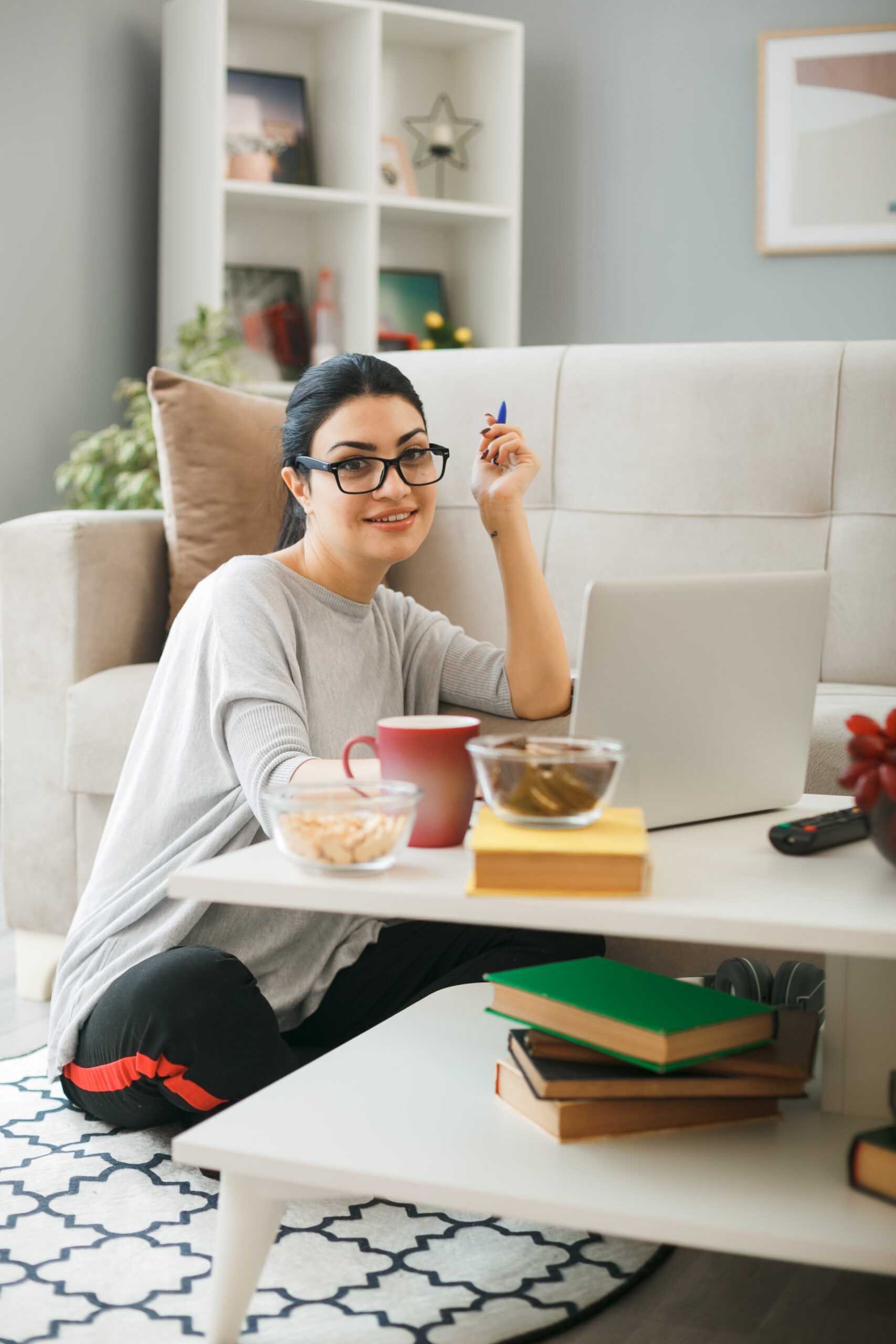 smiling looking camera young girl wearing glasses used laptop sitting on floor behind coffee table in living room